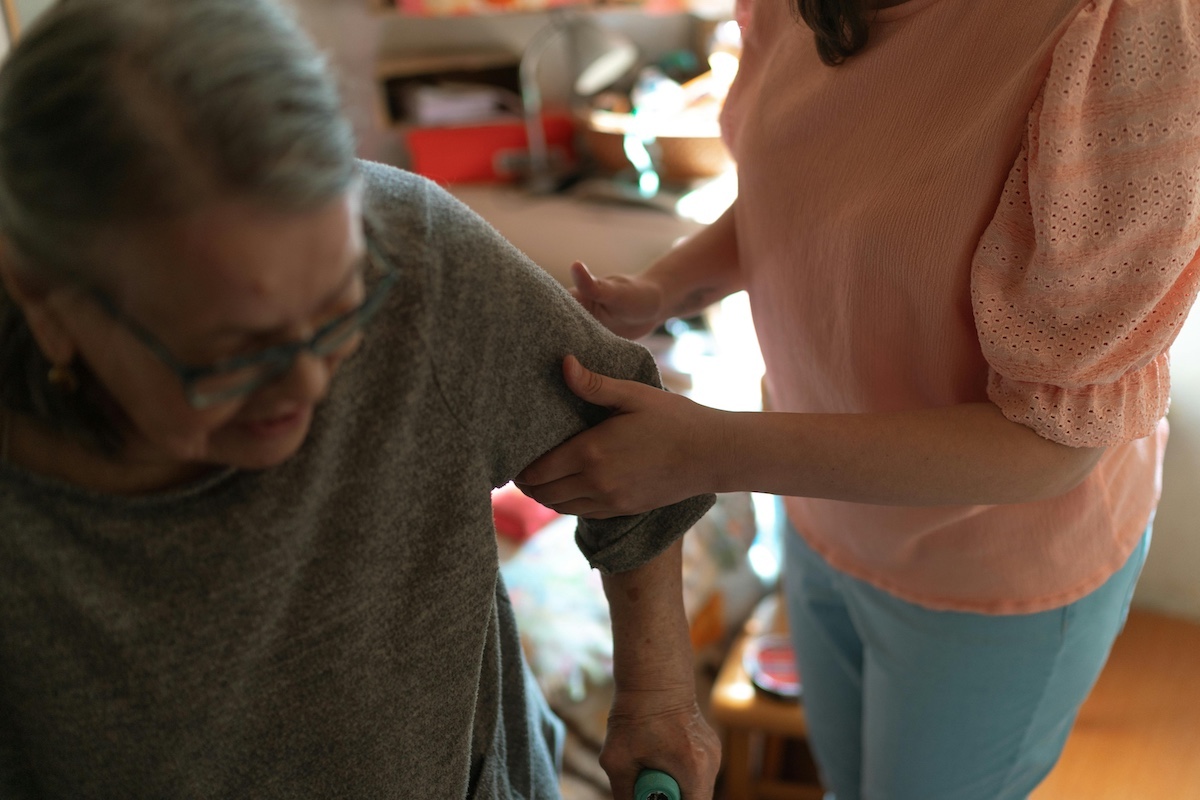 Person in orange shirt helping an older person who is wearing glasses walk